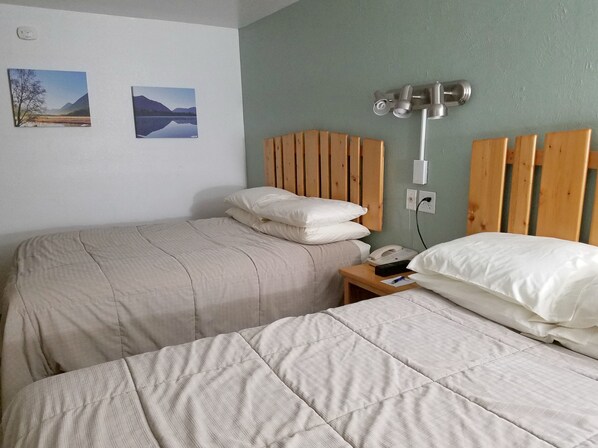 A room with two beds, each with a wooden headboard and white bedding. There are two wall-mounted paintings, a bedside table with a phone, and a power outlet on the wall. - Soldotna Inn (Soldotna)