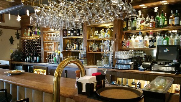 A bar area with a wooden counter, a cash register, and a variety of liquor bottles. - Soldotna Inn (Soldotna)
