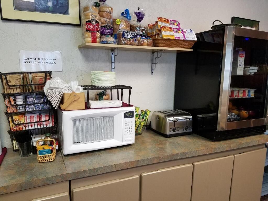 A kitchen counter with a microwave, toaster, and various food items. A sign indicates hot water is located on the coffee maker.