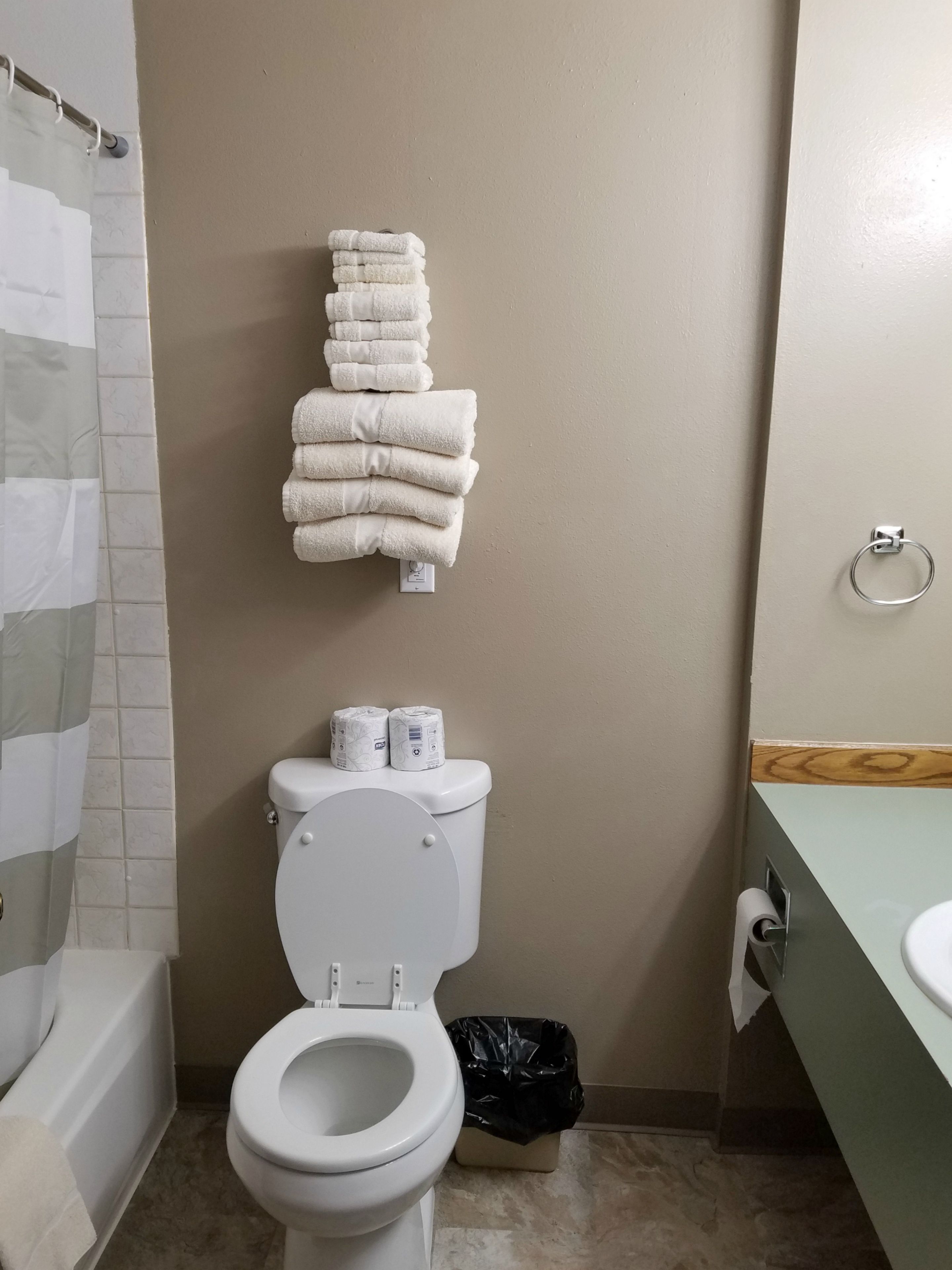 Une salle de bain avec des toilettes, une pile de serviettes blanches, du papier toilette, un sac poubelle noir et un rideau de douche.