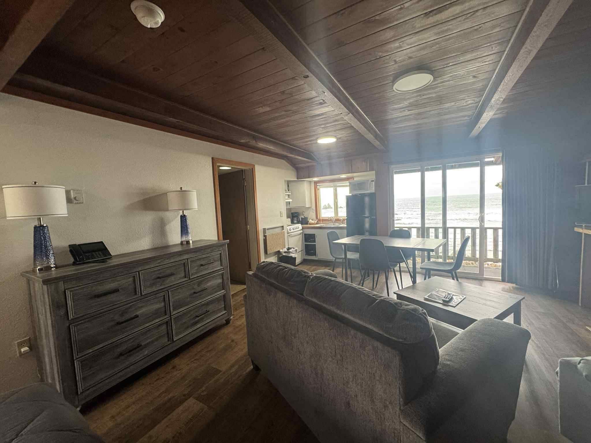 A spacious living area with a wooden ceiling, a sofa, a dining table, and a kitchen area with a view of the sea.