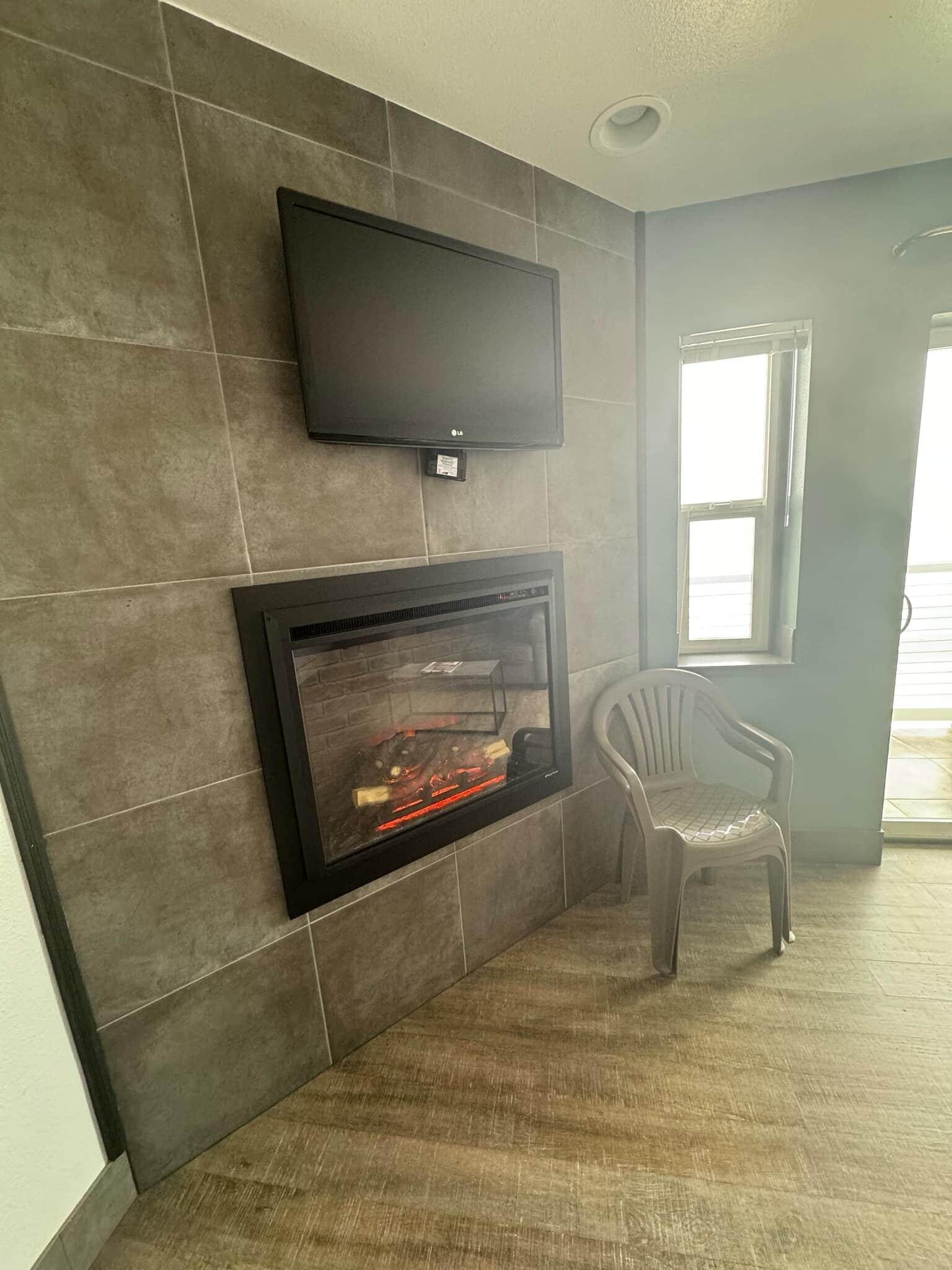Een moderne woonkamer met een flatscreen televisie boven een open haard, een plastic stoel, en een raam dat natuurlijk licht binnenlaat.