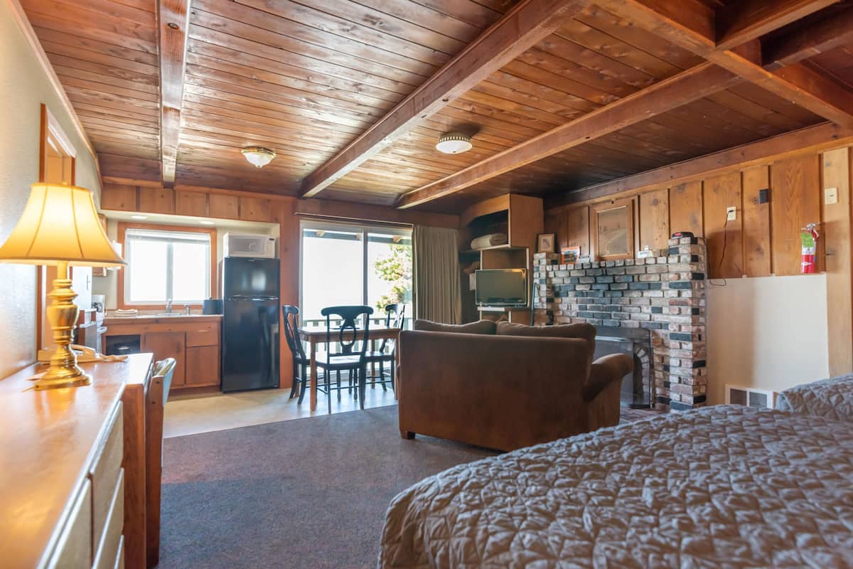 A cozy cabin interior with a wooden ceiling, a dining area, a living room with a sofa, and a kitchen with a brick fireplace.