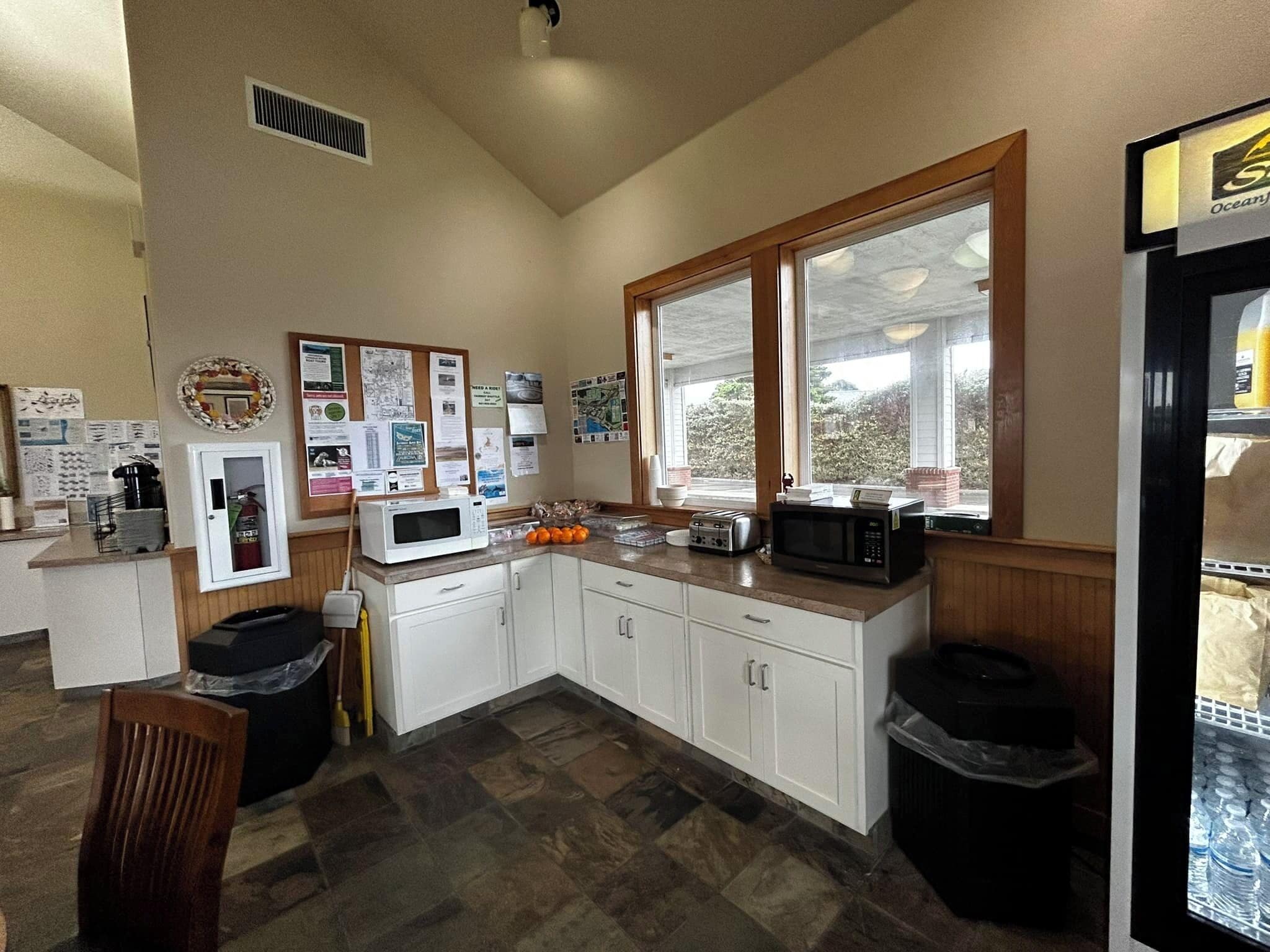 A compact kitchen with white cabinets, a microwave, toaster, and a refrigerator. There are bulletin boards on the wall and a window providing natural light.