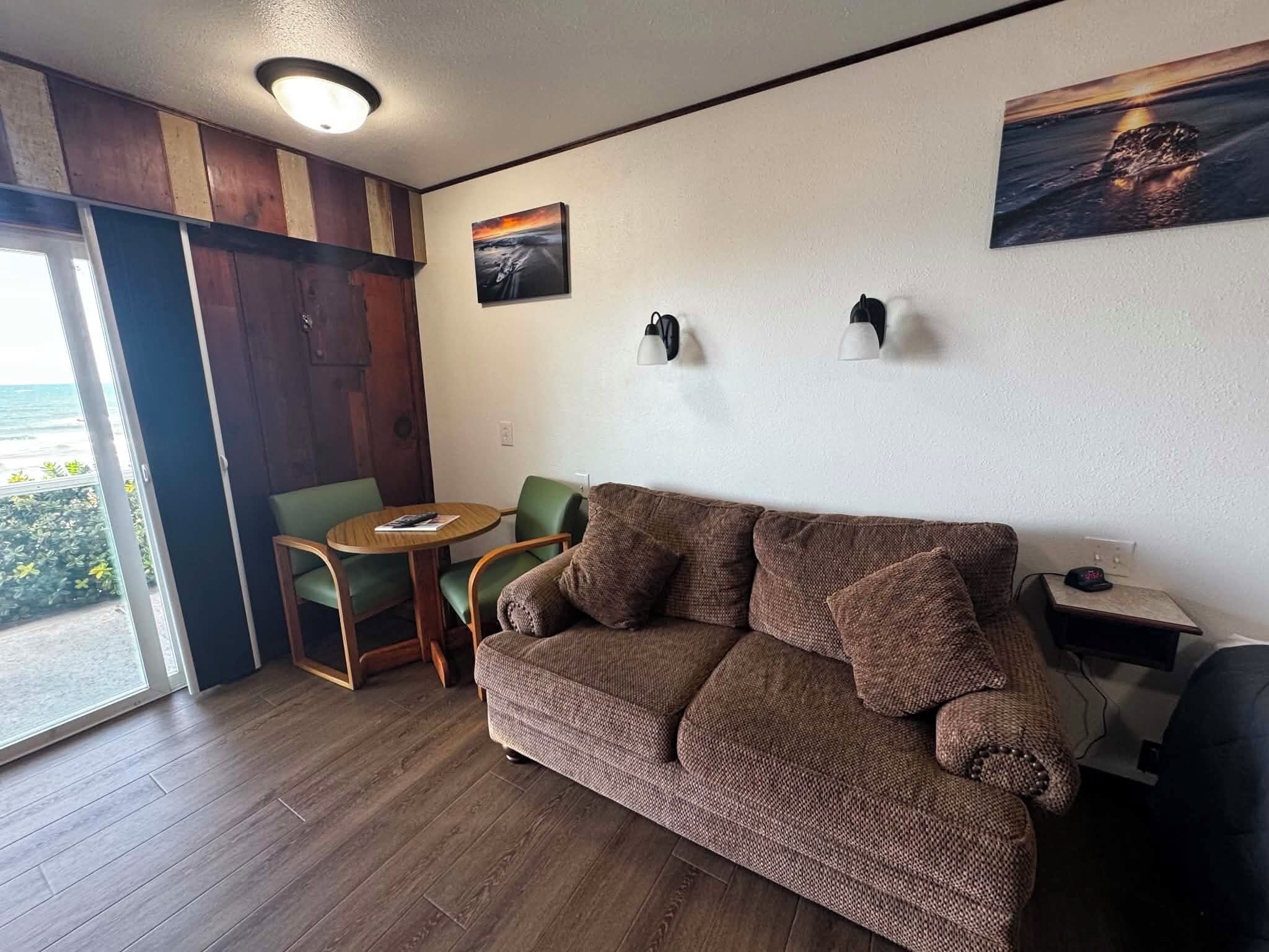 A cozy living area with a brown sofa, a small round table, and two chairs. There is a view of the outdoors through a glass door.