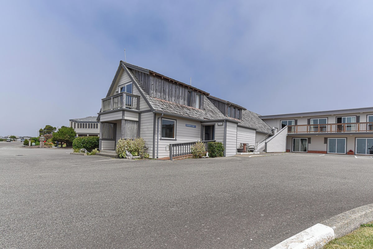 A two-story building with a sloped roof, a front entrance, and a small porch area with plants.