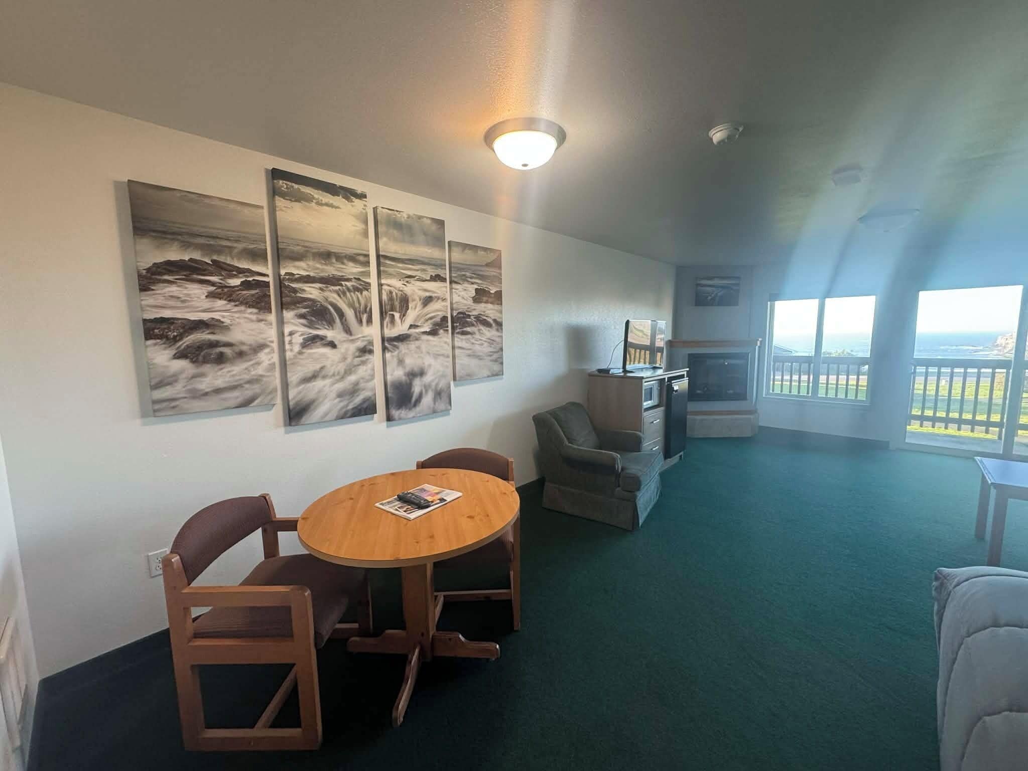 A room with a round wooden table, two chairs, a television, and a fireplace. There are three large paintings on the wall depicting ocean scenes.