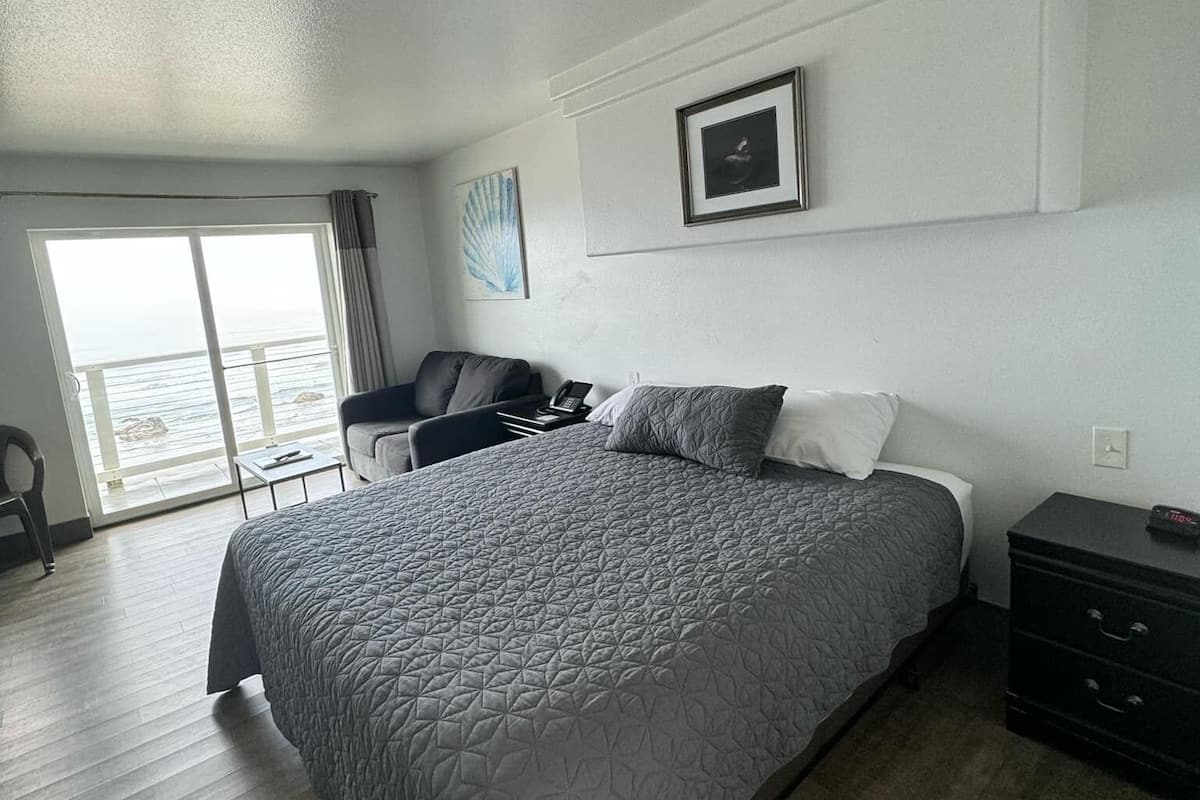 A modern hotel room with a large bed, a desk, and a chair. There is a window with a view of the sea.