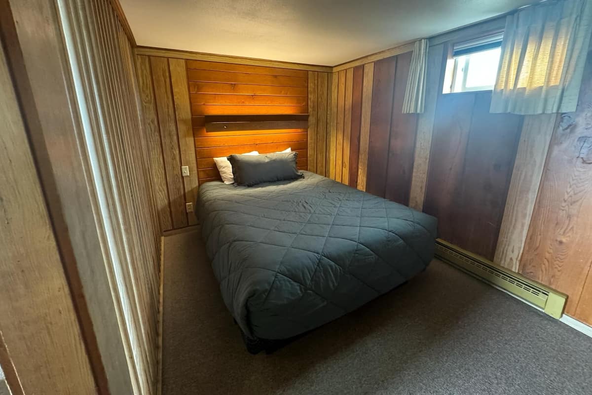 A bedroom with a wooden headboard, a bed with a dark quilt, and a window with curtains.