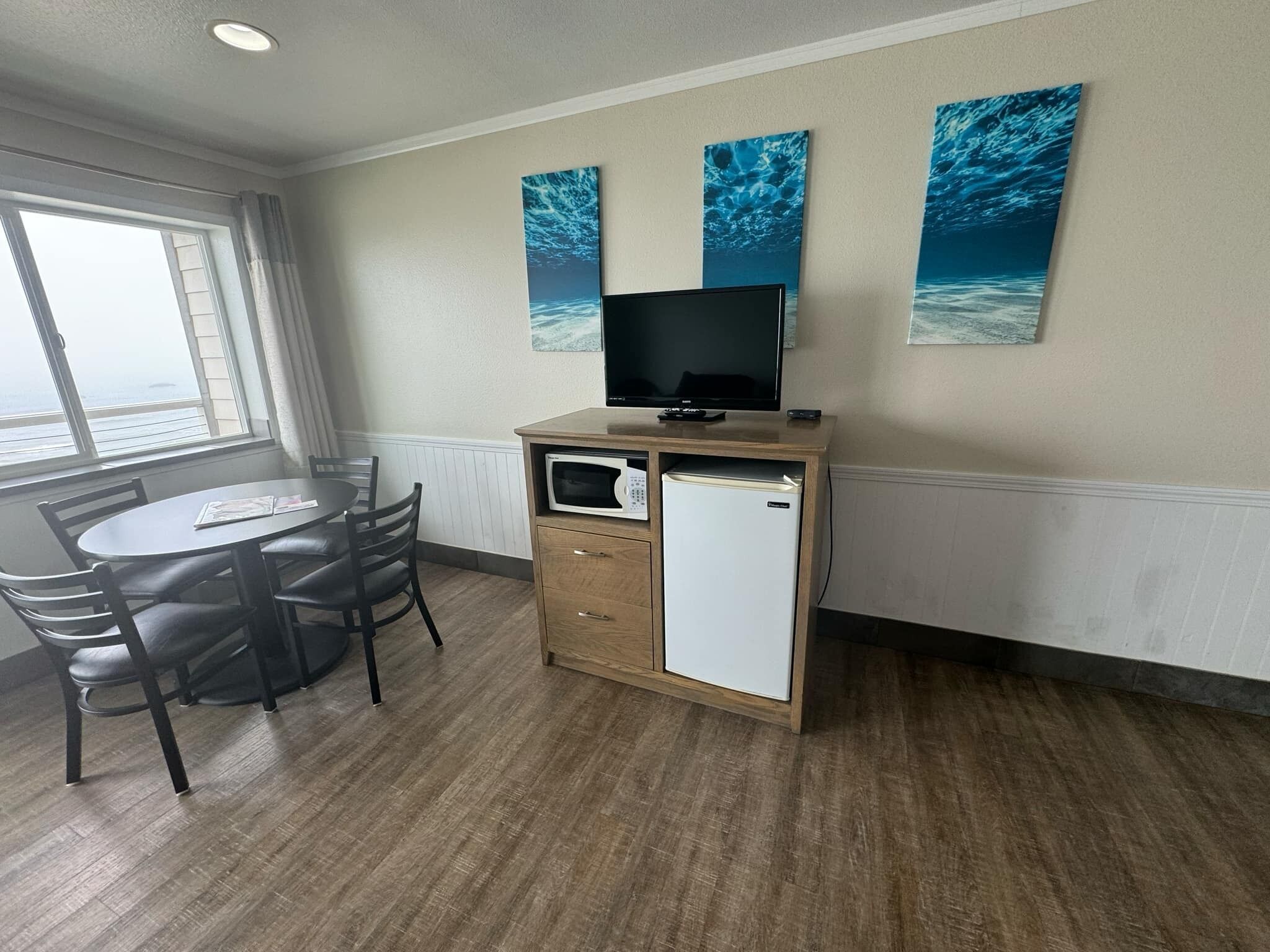 A compact room with a wooden desk, a television, a small refrigerator, and a dining table with chairs. There are three wall-mounted paintings depicting ocean scenes.