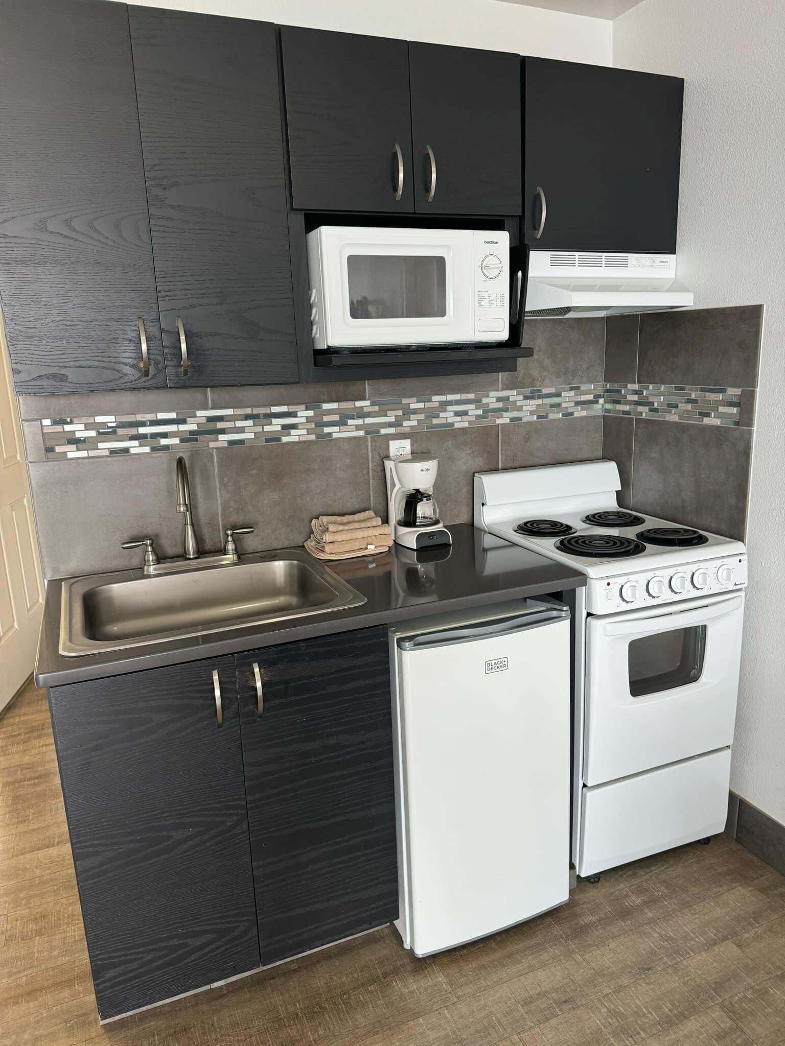 A compact kitchen with dark cabinets, a white microwave, a coffee maker, a stove, and a small refrigerator.