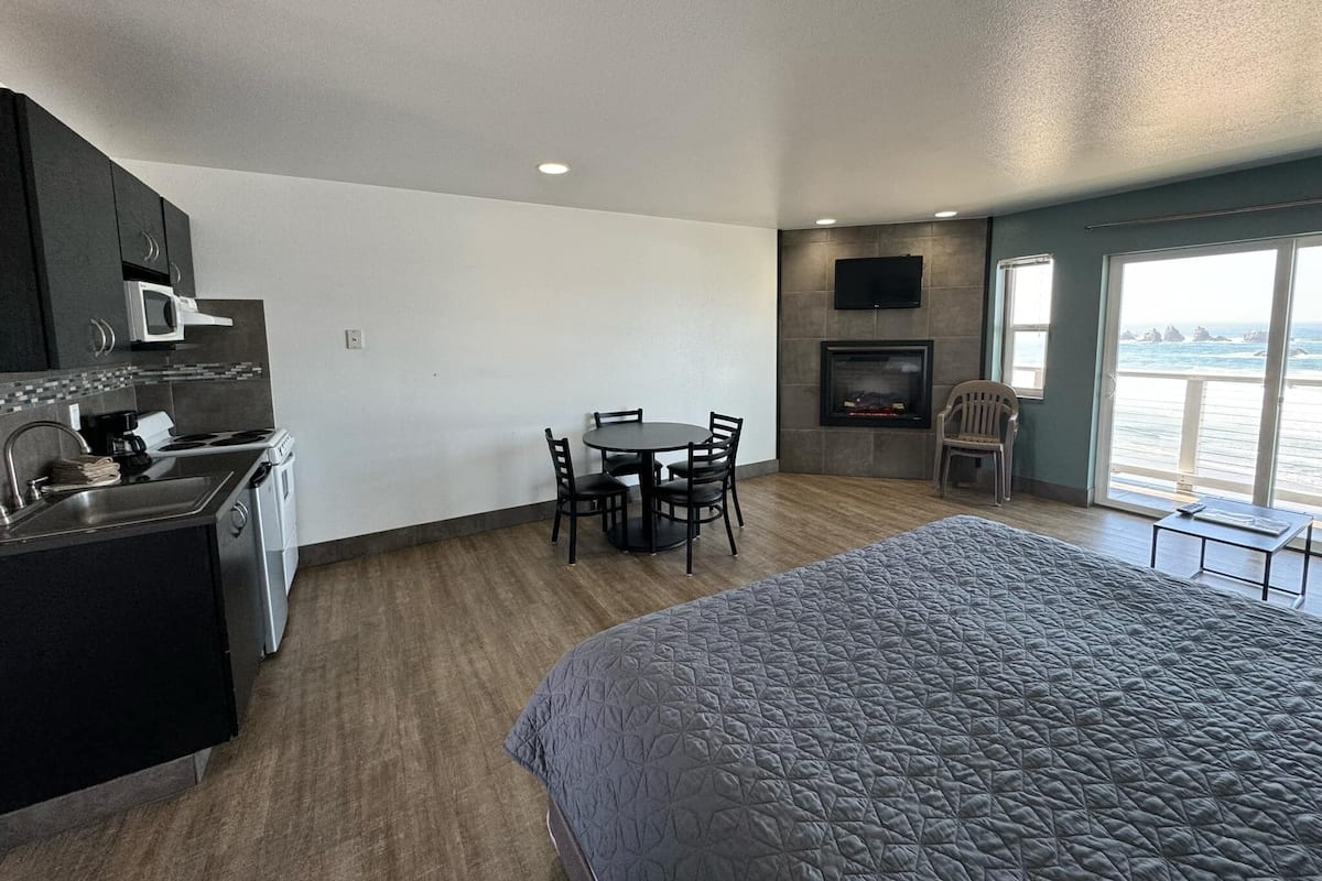 A compact hotel room with a kitchenette, a dining area, a bed, a fireplace, and a large window with a view.