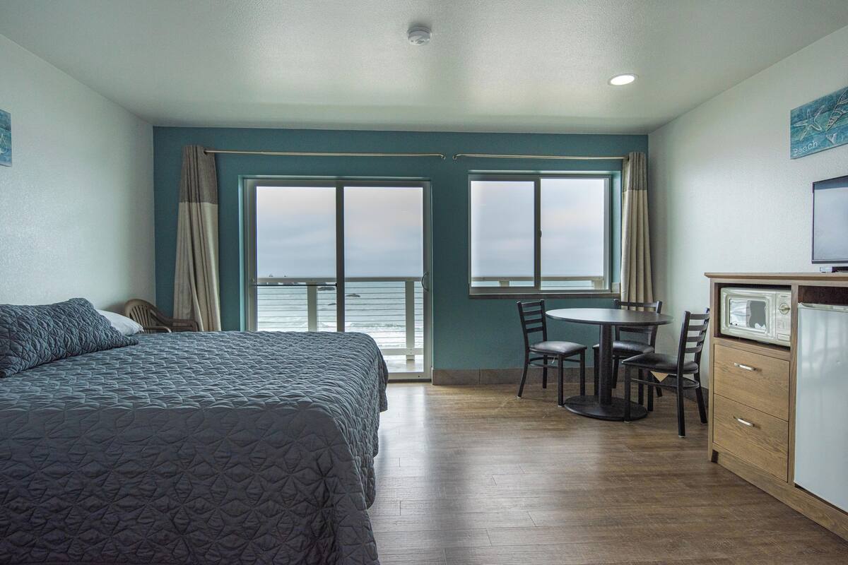 A hotel room with a bed, a dining table with chairs, a TV, and a view of the sea through large windows.