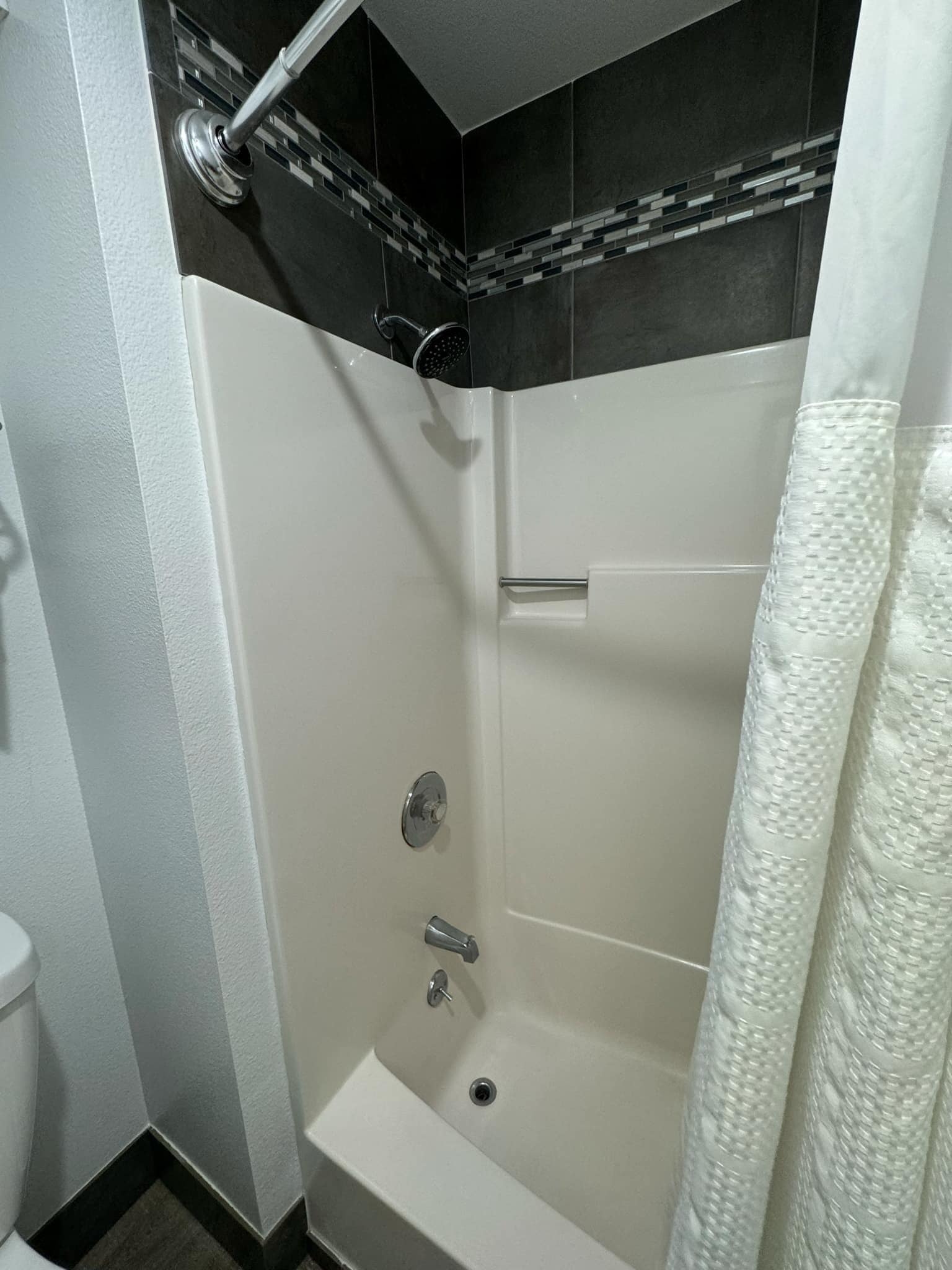 A shower with a handheld showerhead, a white bathtub, and a tiled wall.