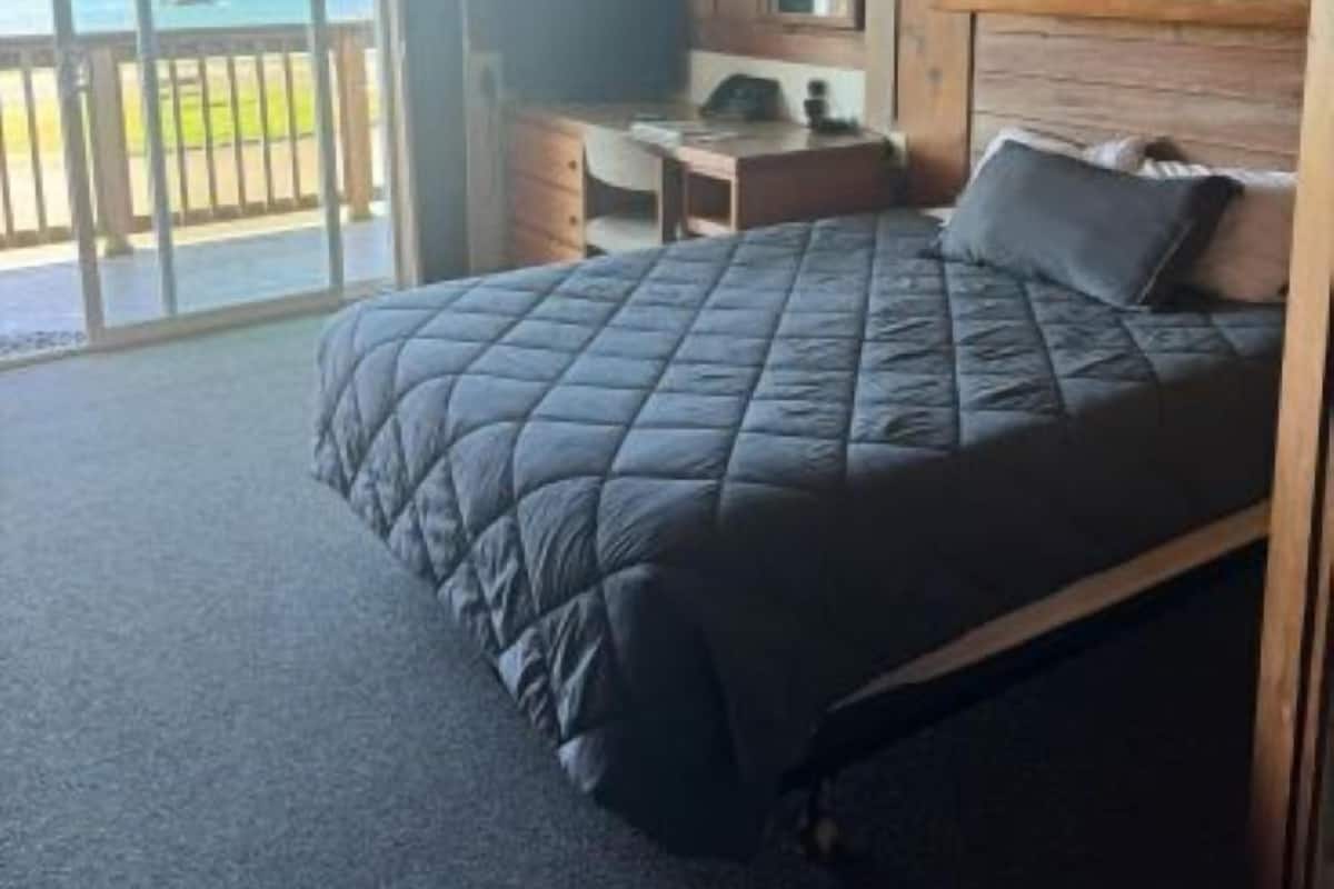 A bedroom with a bed, a desk, a chair, and a view of the ocean through a sliding door.