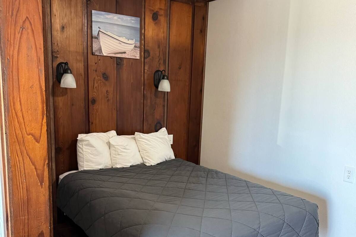 A bedroom with a wooden headboard, a gray quilted bedspread, two white pillows, and two wall-mounted lamps.