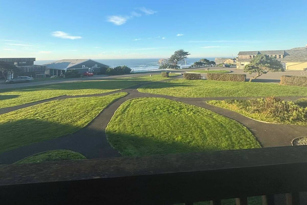 A view from a balcony overlooking a green landscape with pathways, a body of water, and buildings in the distance.
