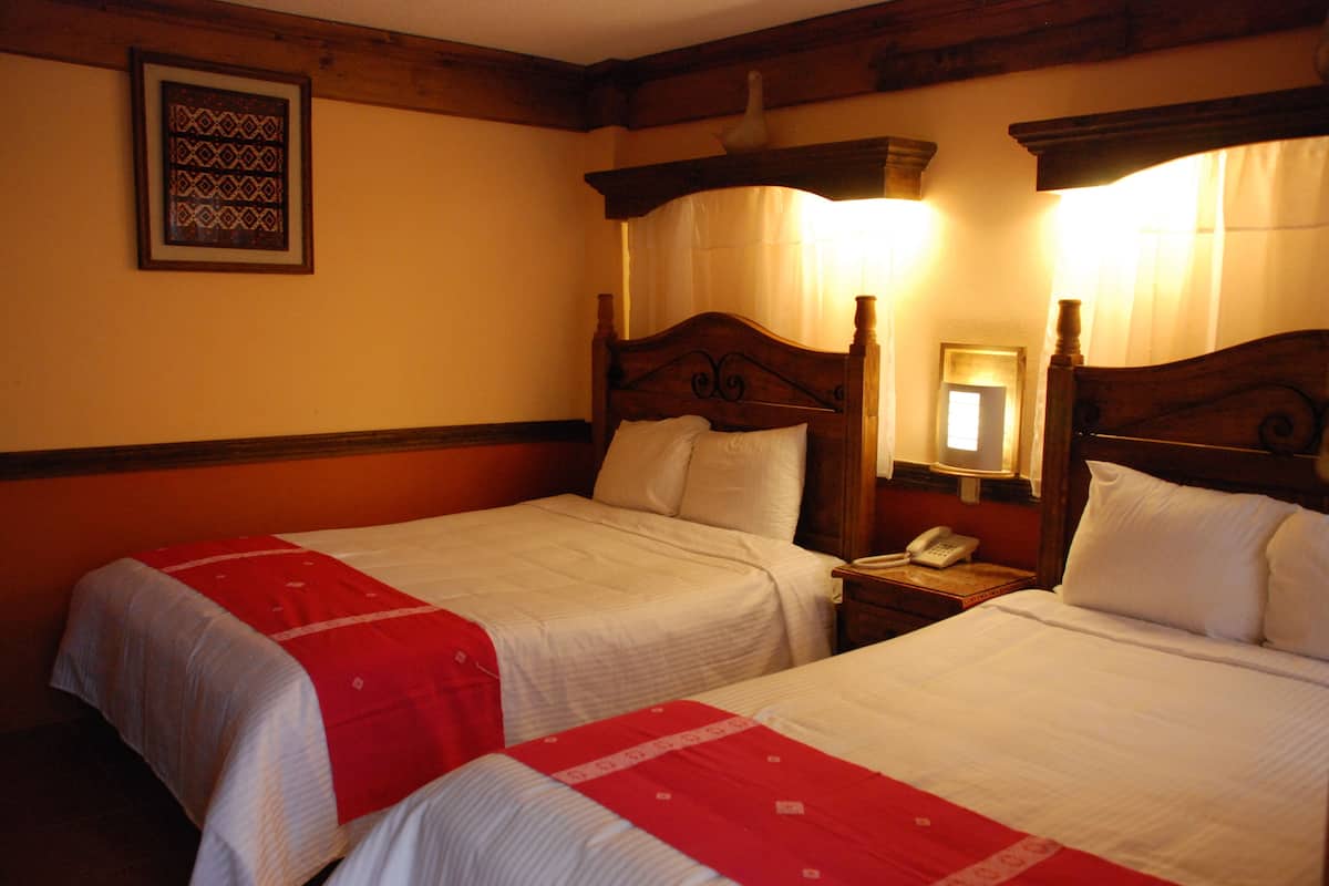 A hotel room with two single beds, wooden headboards, a bedside table with a lamp, and a framed picture on the wall.