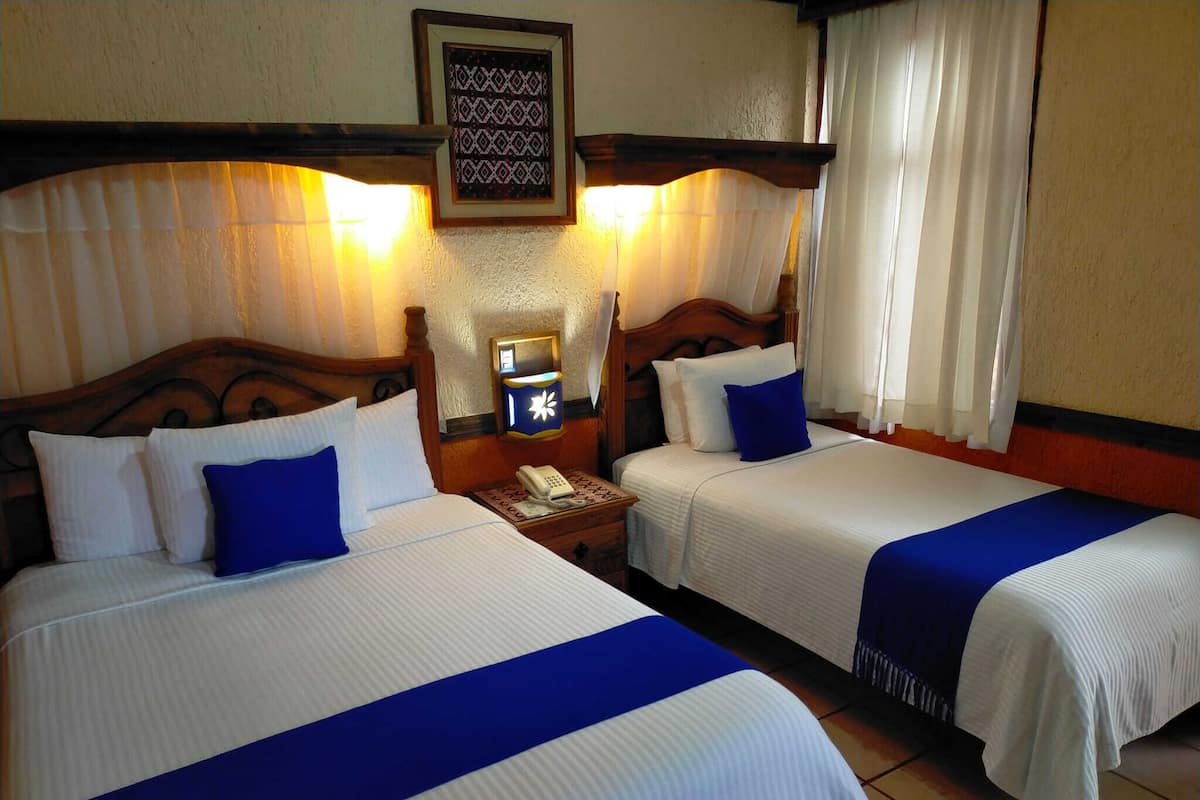 A hotel room with two beds, each with white bedding and blue accents. There is a nightstand with a phone and a wall-mounted light between the beds.