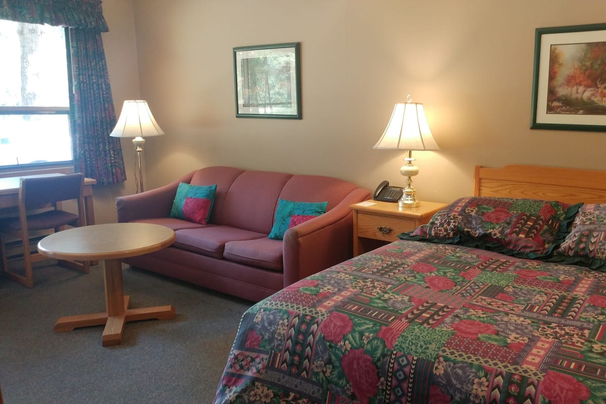 Habitación de hotel con cama, sofá, una mesita, dos lámparas y dos cuadros en la pared.