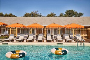 Un área junto a la piscina con sillas de descanso y sombrillas naranjas, una piscina azul cristalina y una valla de madera al fondo.