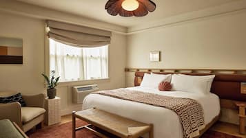 A bedroom with a bed, a bench, a chair, a window with curtains, a wall-mounted air conditioner, and a ceiling light fixture.