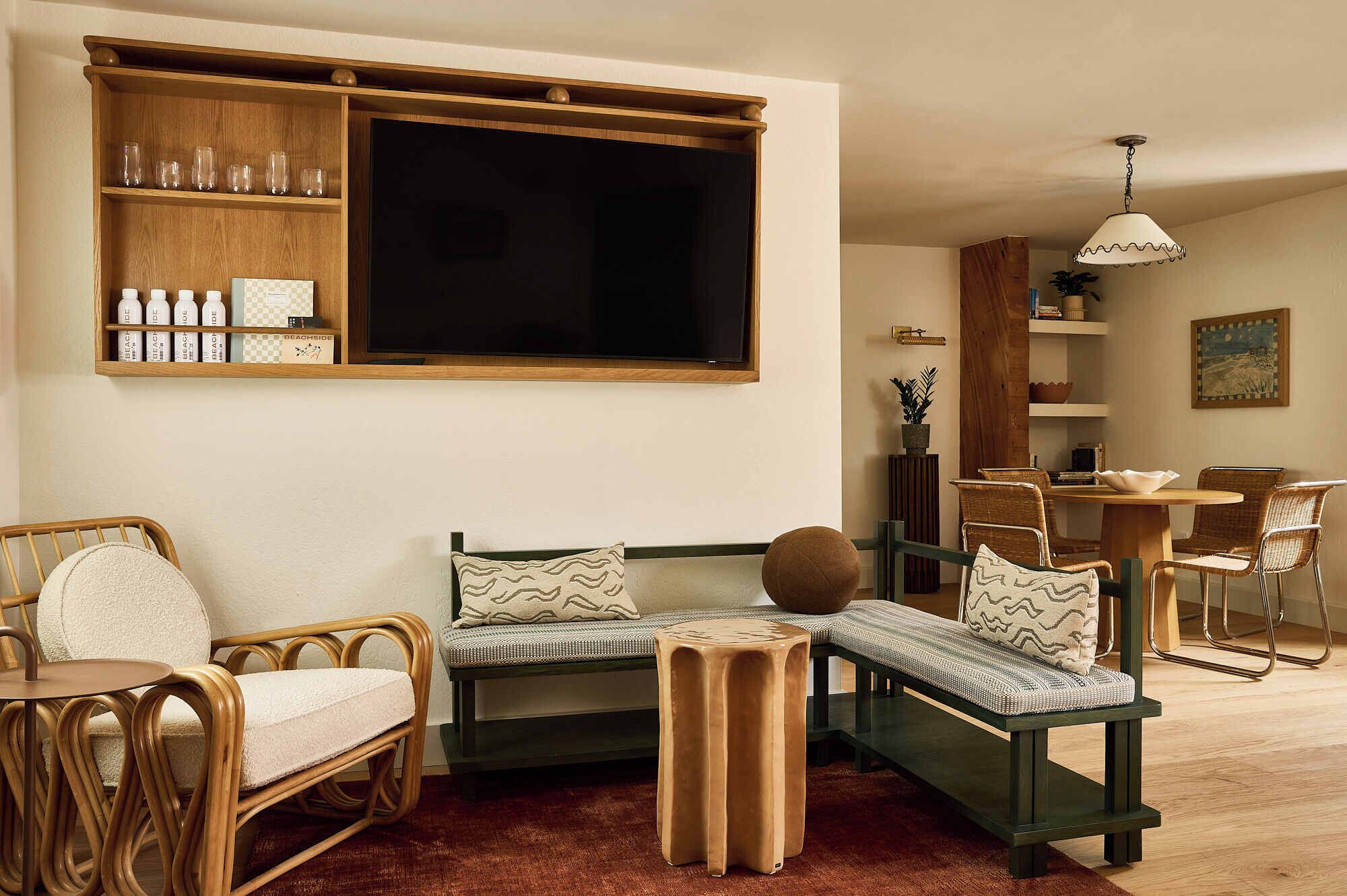 Ein gemütliches Wohnzimmer mit einem großen Fernseher, Korbmöbeln, einem Holzregal mit Flaschen und einem Essbereich mit Tisch und Stühlen.