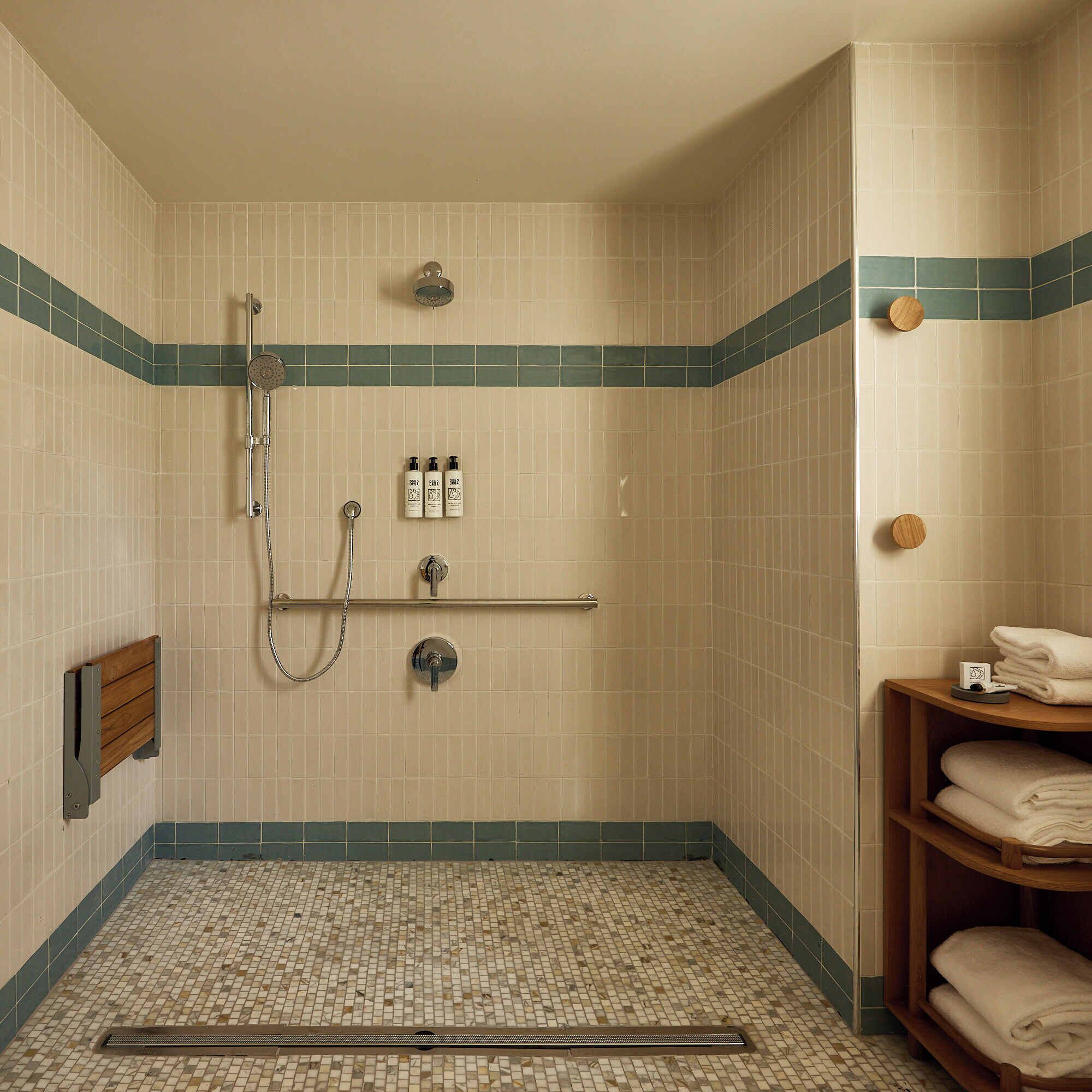 Ein kleines, mit Fliesen ausgelegtes Duschbad mit Duschkopf, Handbrause und einem Brausensauger. An der Wand befinden sich drei Flaschen Shampoo und Conditioner, und ein Handtuchhalter mit gefalteten Handtüchern.