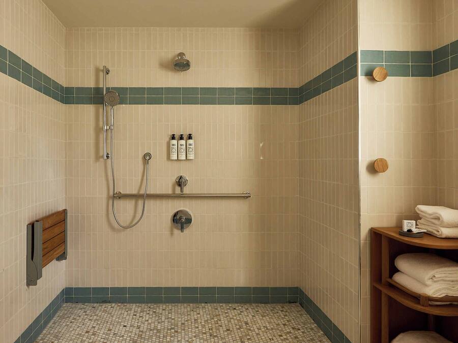 A small, tiled shower room with a showerhead, handheld shower, and a handheld shower attachment. There are three bottles of shampoo and conditioner on the wall, and a towel rack with folded towels.