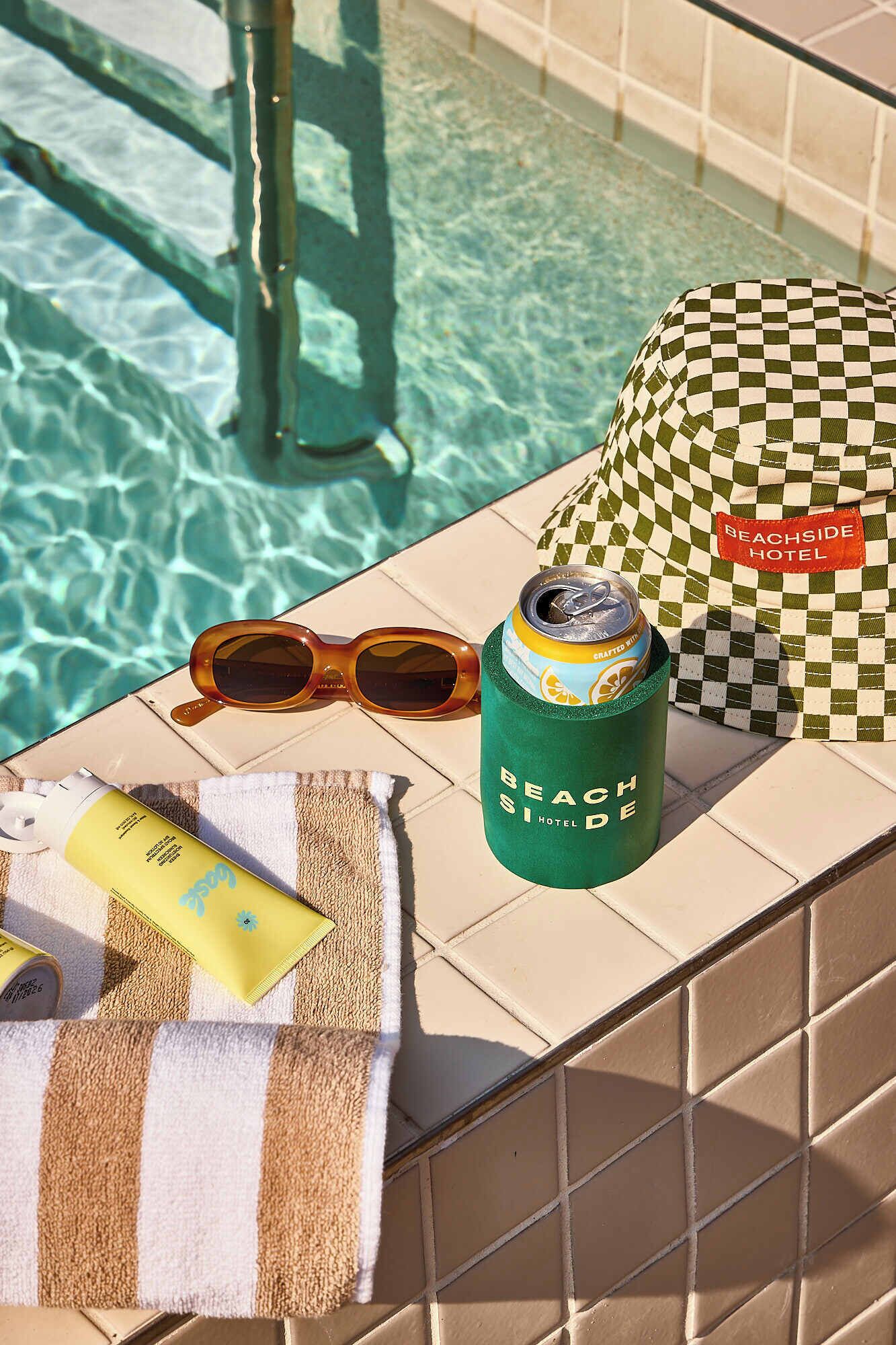 A poolside setting with a towel, sunglasses, a can of beverage, and a checkered bag, indicating a relaxing environment.