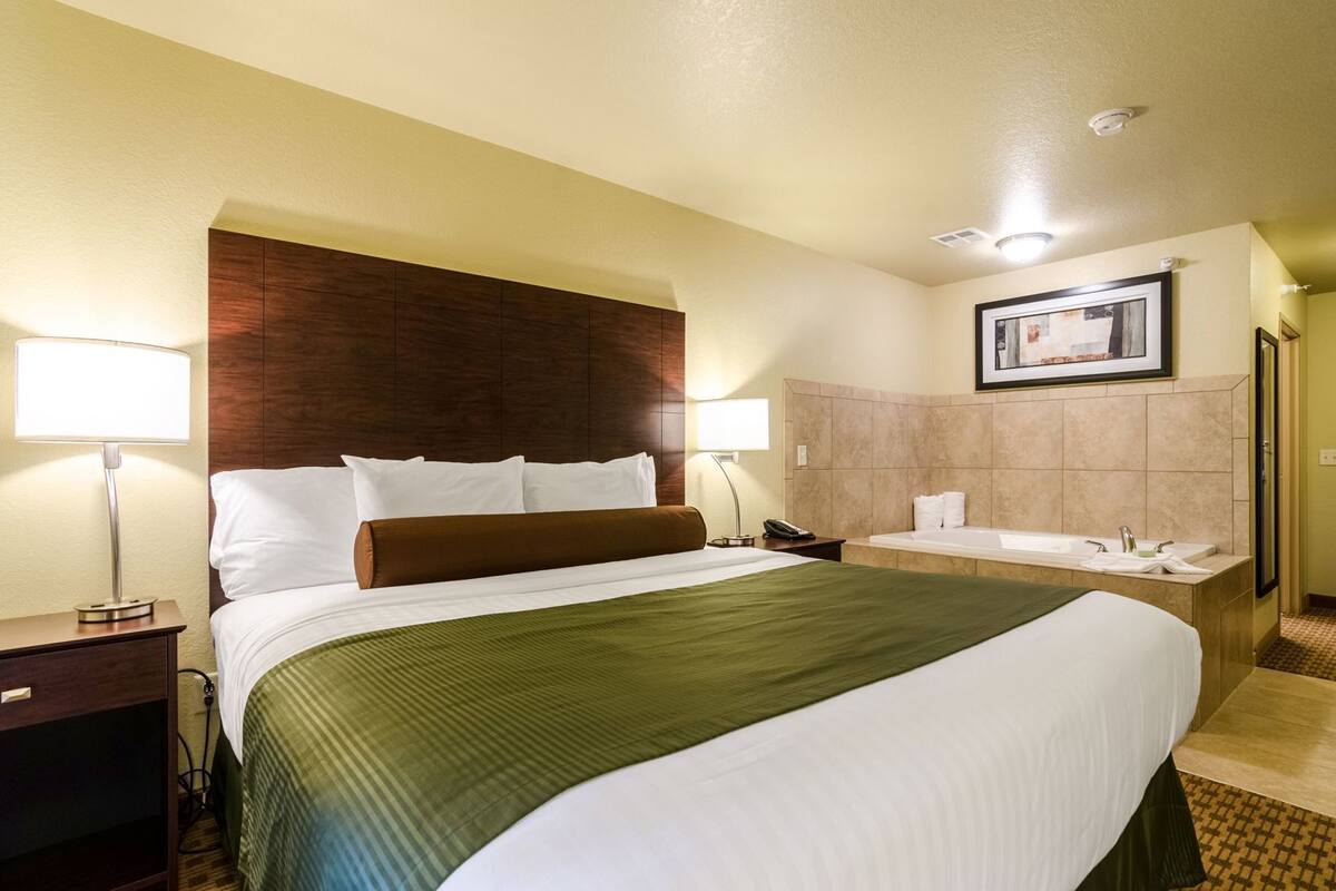 A hotel room with a bed, bedside lamps, a nightstand, a bathroom with a bathtub, and a framed picture on the wall.