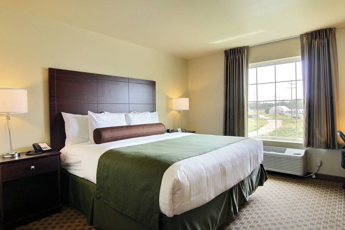 A hotel room with a large bed, a nightstand with a lamp, a window with curtains, and an air conditioning unit.