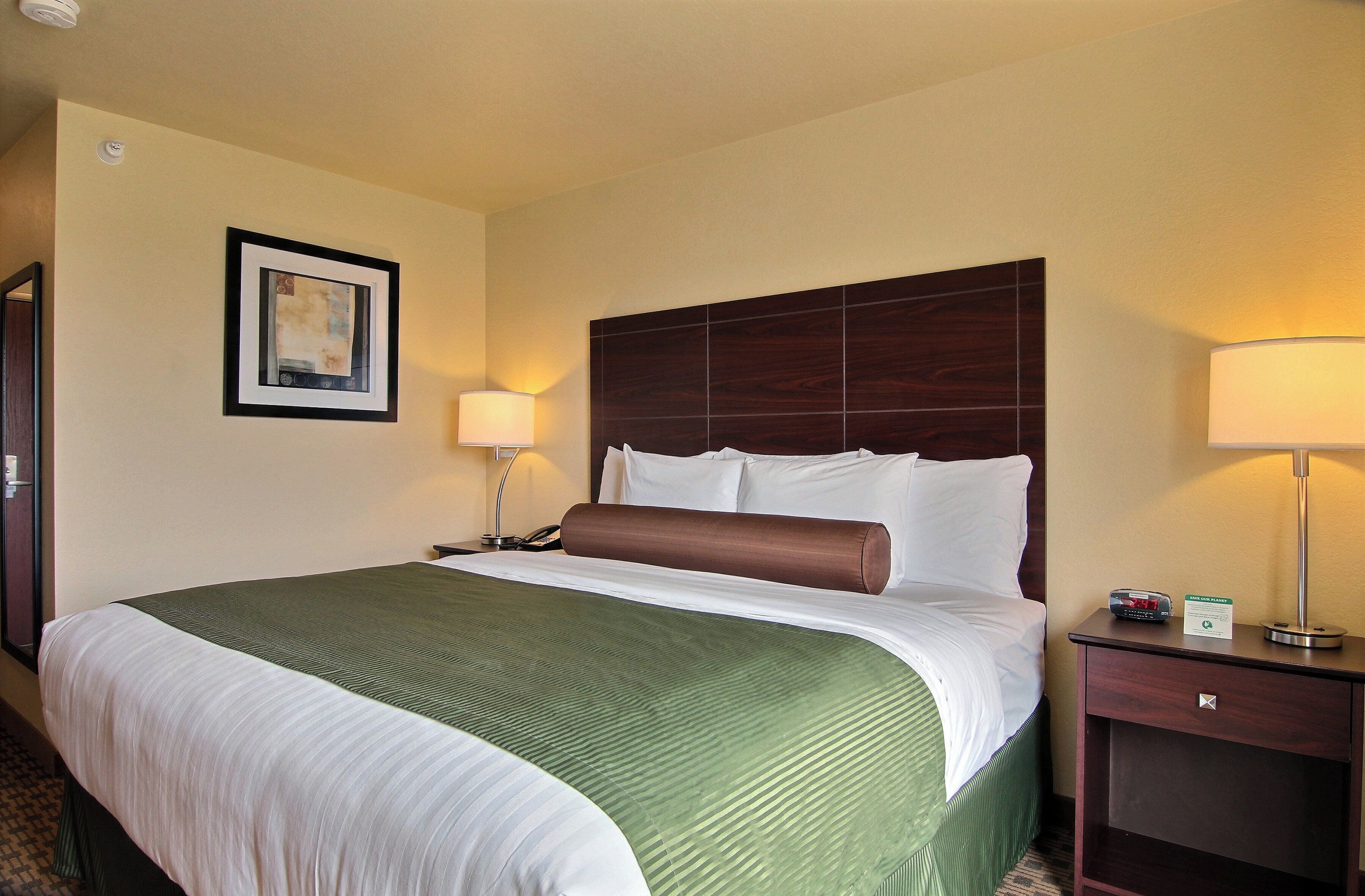 A hotel room with a large bed, a wooden headboard, a bedside table with a lamp, and a framed picture on the wall.