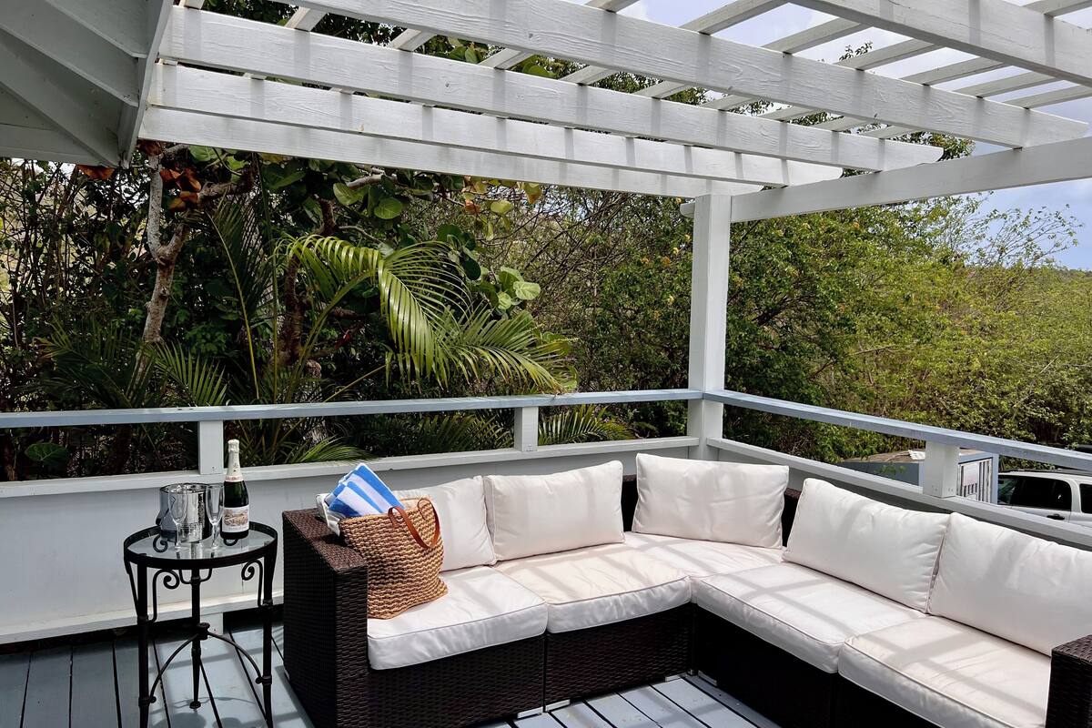 A covered outdoor seating area with a white pergola, wicker furniture, and a small table with a bottle and glasses.