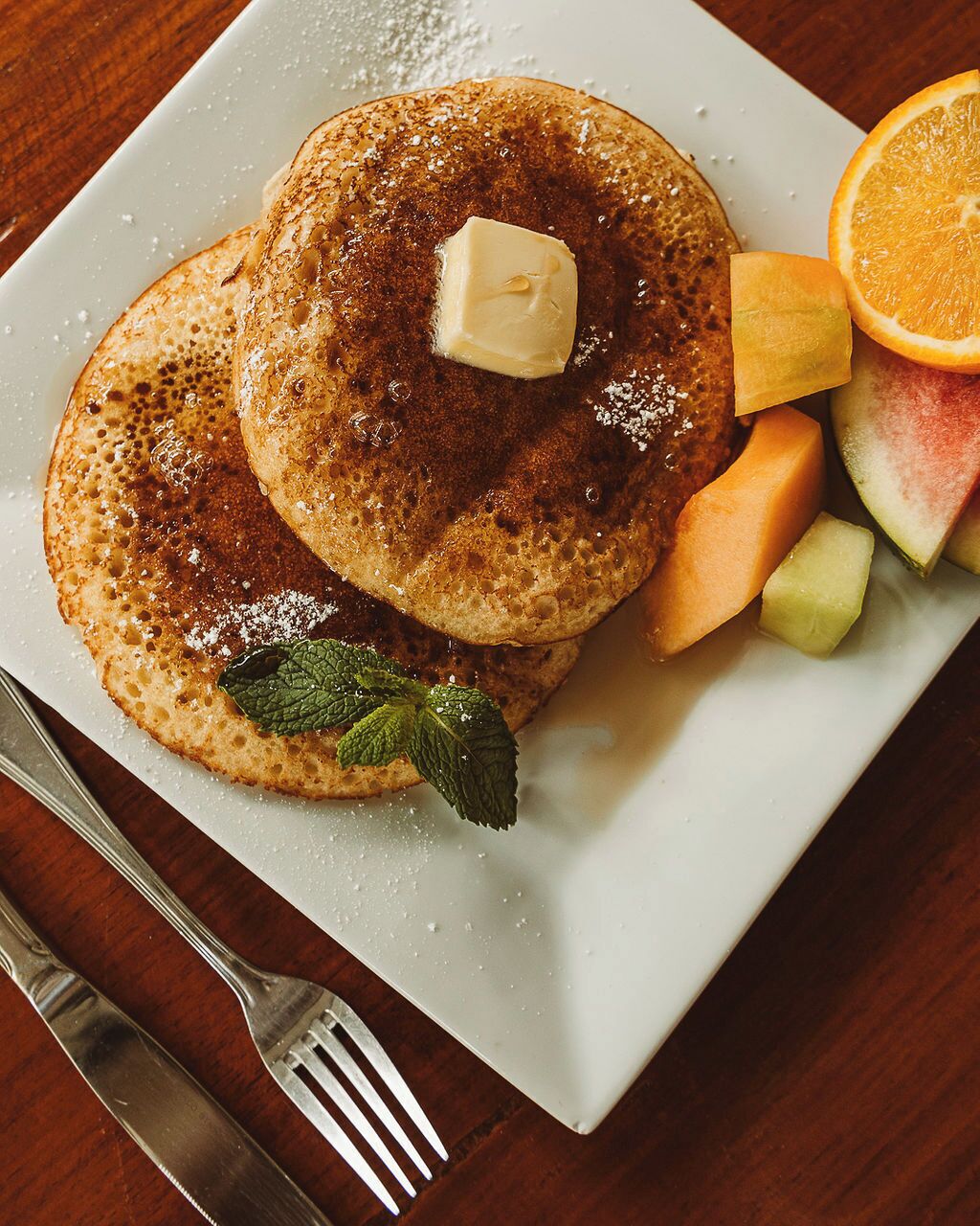 A plate with pancakes topped with butter and syrup, accompanied by a slice of orange and melon pieces.