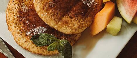 A plate with pancakes topped with butter and syrup, accompanied by a slice of orange and melon pieces.