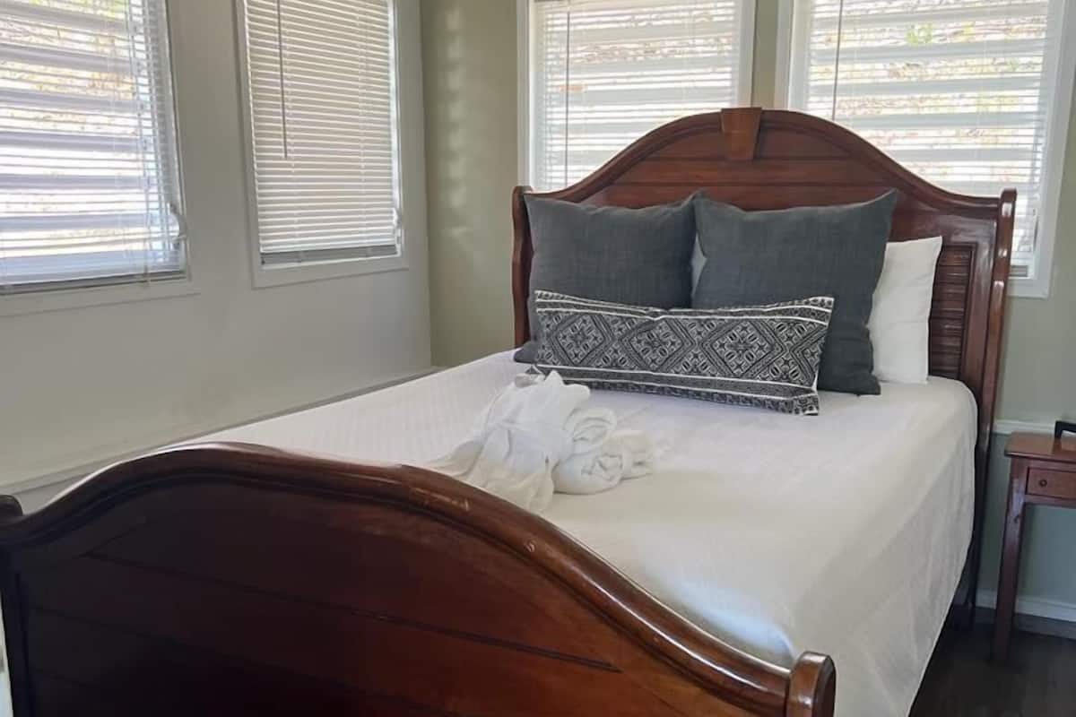 A bedroom with a wooden bed, white bedding, and three windows with blinds.