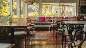 An indoor seating area with wicker furniture, wooden flooring, and large windows offering a view of greenery.