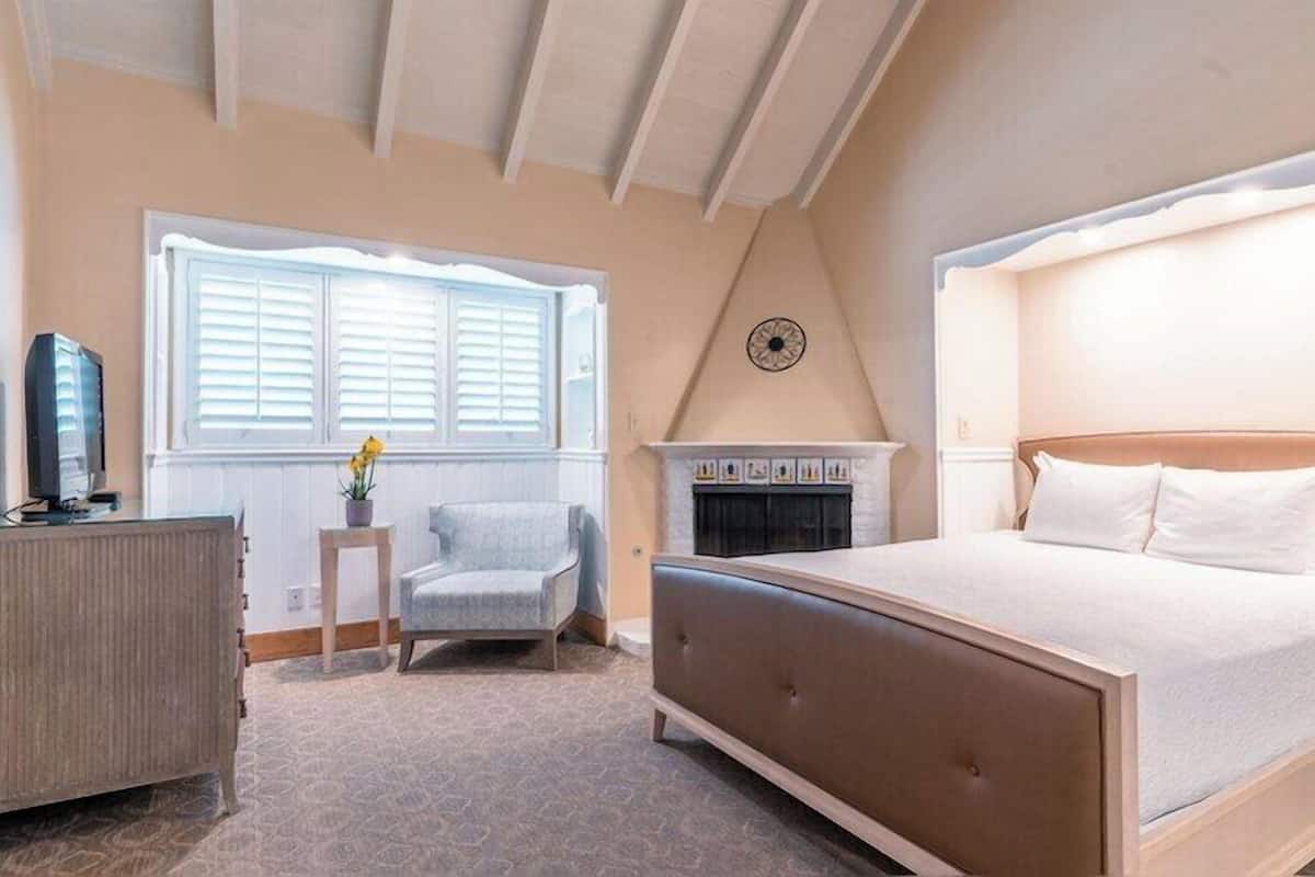 A bedroom with a large bed, a chair, a dresser, a fireplace, and a window with blinds.