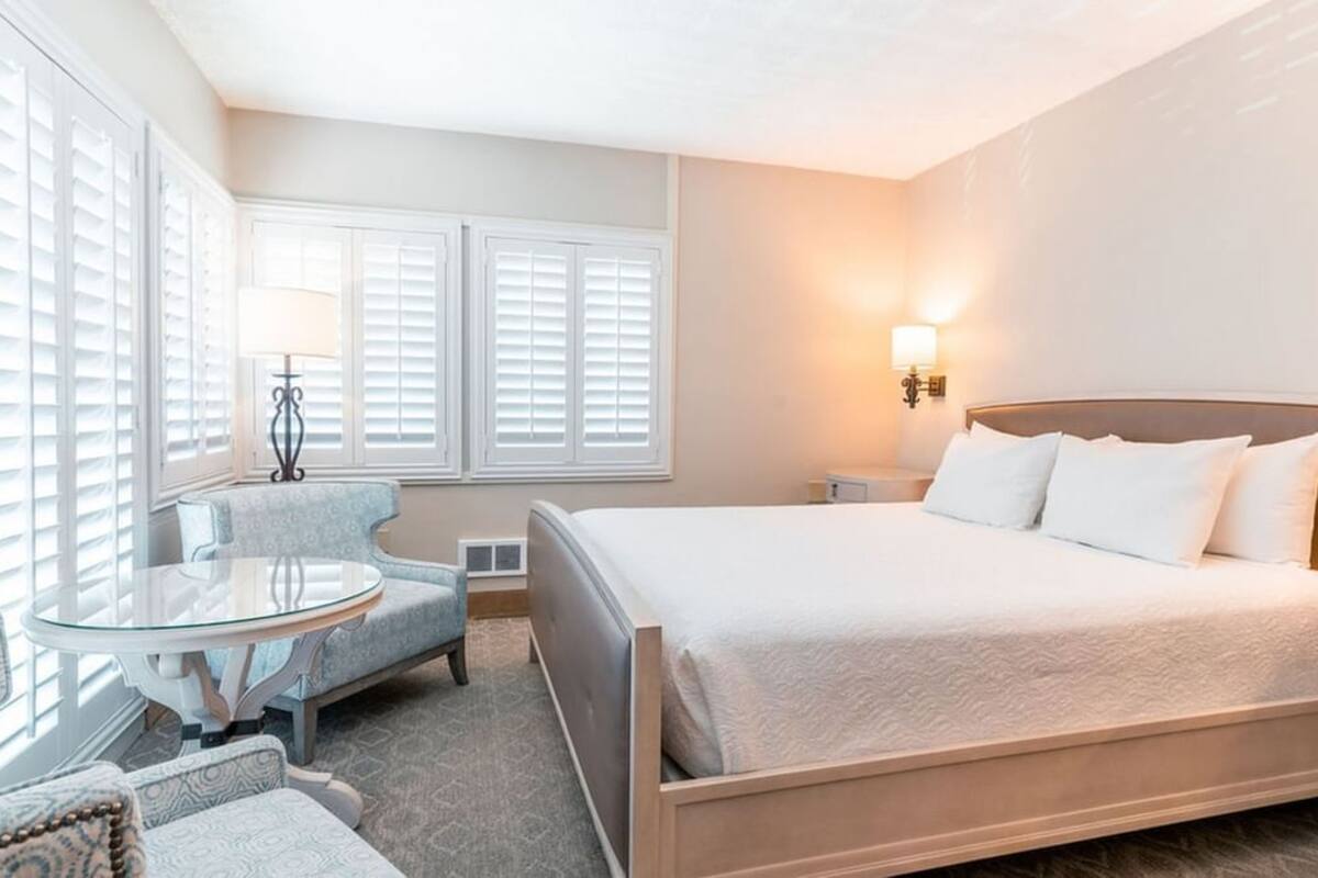 A hotel room with a large bed, a round glass table, two armchairs, and a nightstand with a lamp.