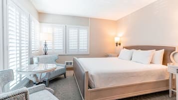 A hotel room with a large bed, a round glass table, two armchairs, and a nightstand with a lamp.