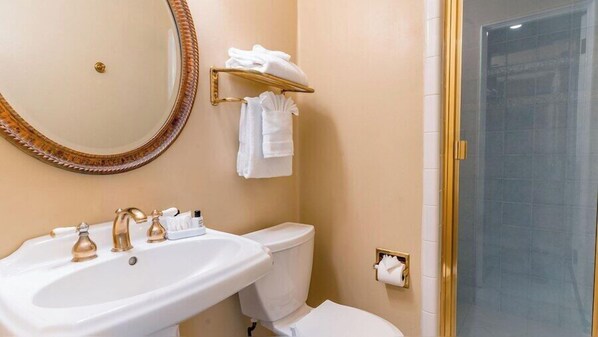 A bathroom with a white sink, gold faucets, a toilet, a towel rack with towels, a mirror, and a shower door.
