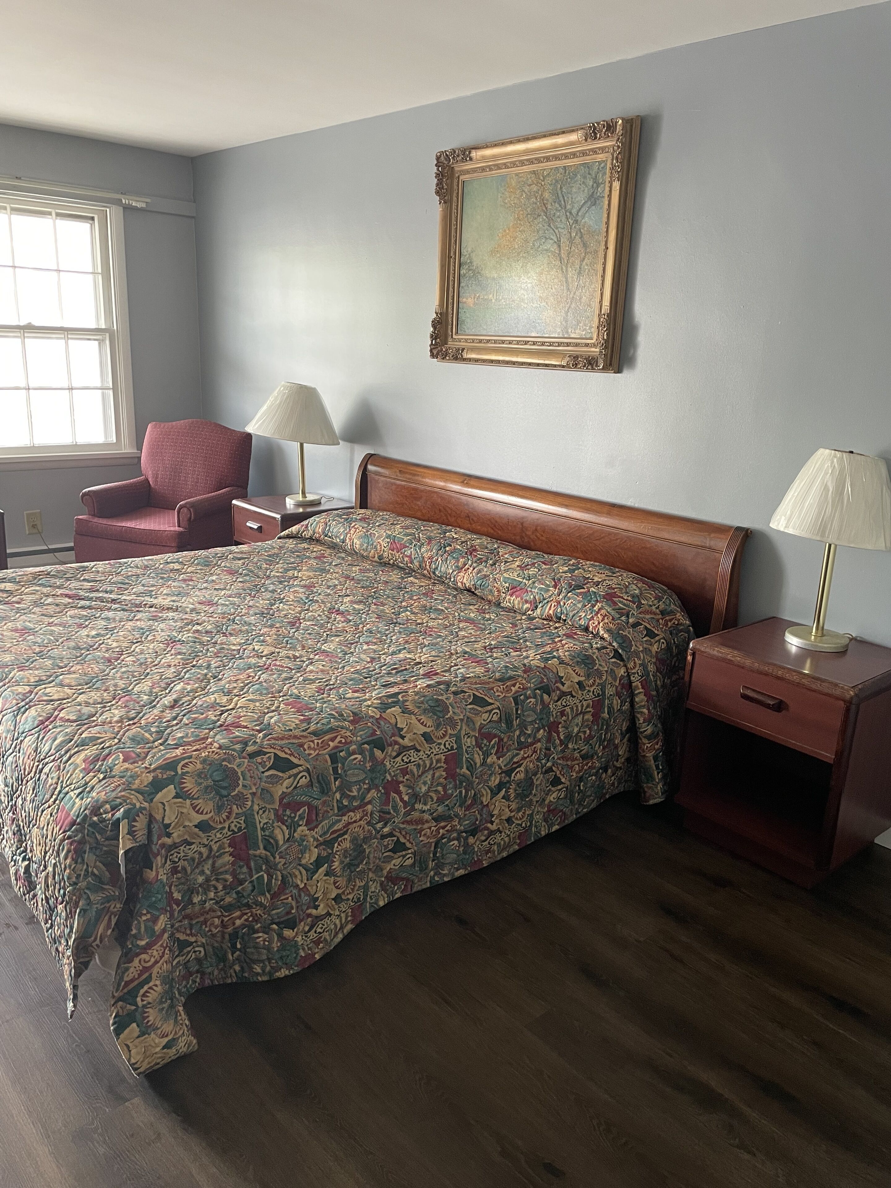 A bedroom with a large bed, a chair, two bedside tables, a window, and a framed picture on the wall.