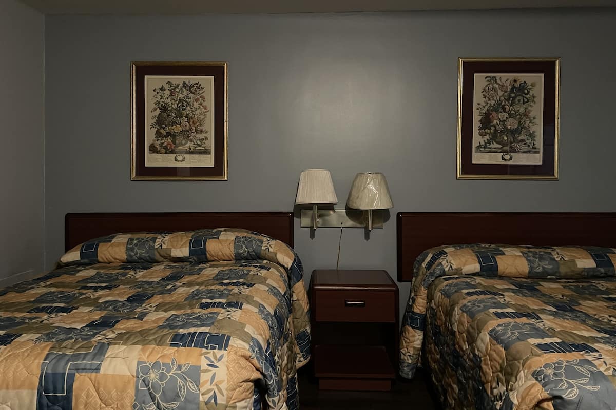 A hotel room with two beds, each with a quilted bedspread, a nightstand with a lamp, and two framed floral pictures on the wall.