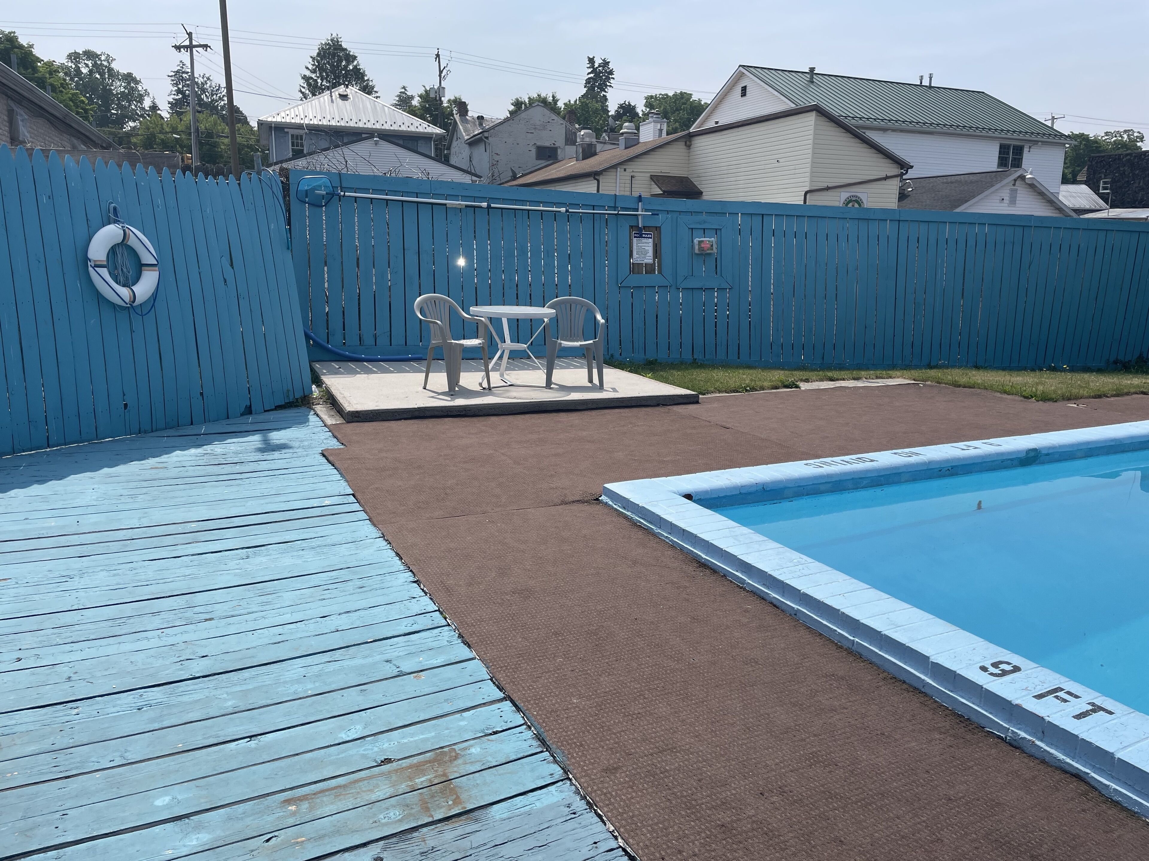 Un patio trasero con piscina, cerca azul y zona de patio con sillas y mesa.