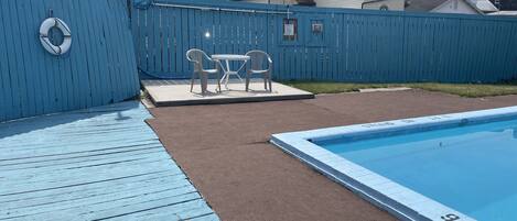 A backyard with a pool, a blue fence, and a patio area with chairs and a table.