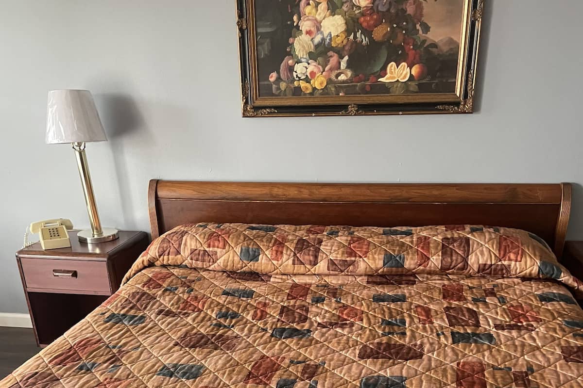 A bed with a quilted bedspread featuring a geometric pattern, a wooden headboard, a nightstand with a lamp, and a framed floral painting on the wall.