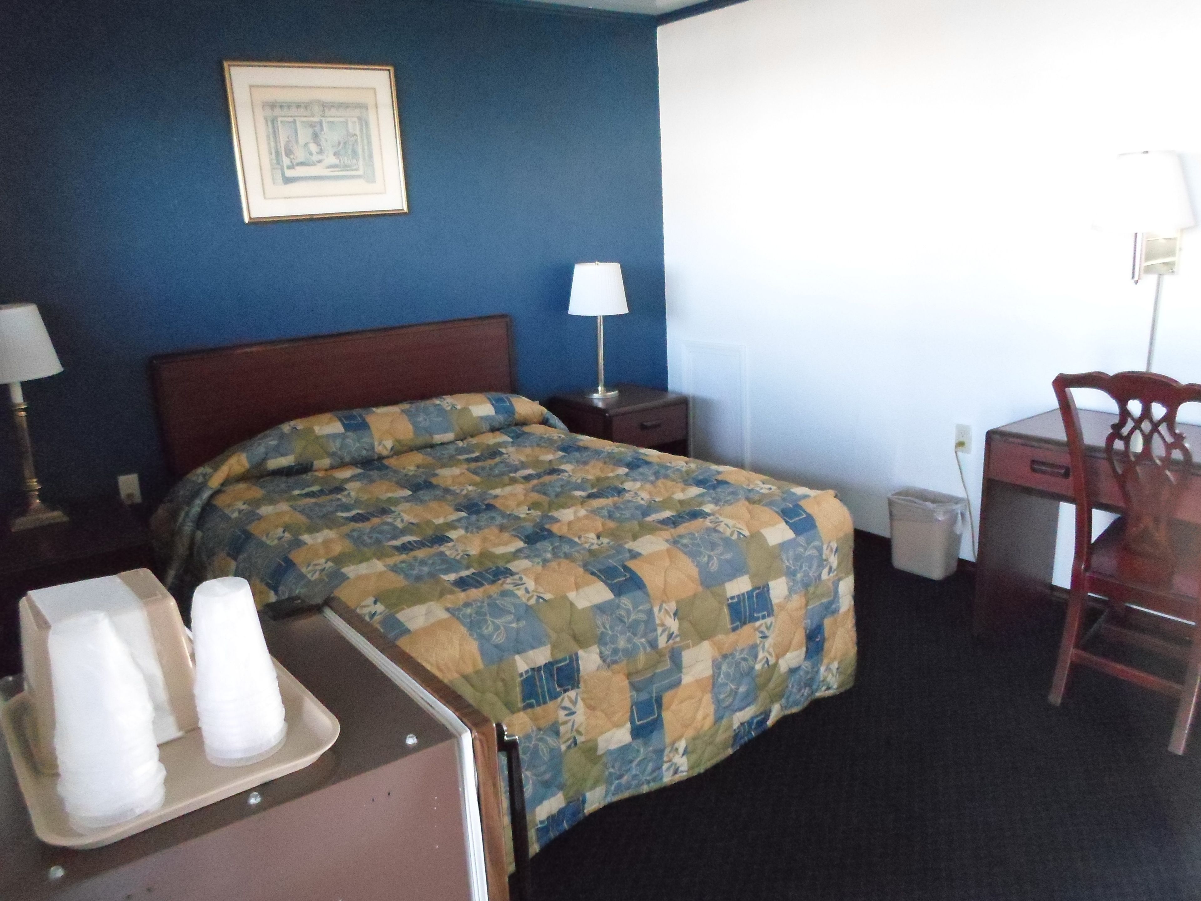 A hotel room with a bed, a desk, a chair, a lamp, and a picture on the wall.