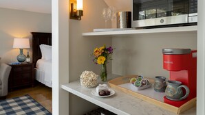 Habitación de hotel con cama, mesita de noche, lámpara, microondas, cafetera y una bandeja con aperitivos y un jarrón con flores.