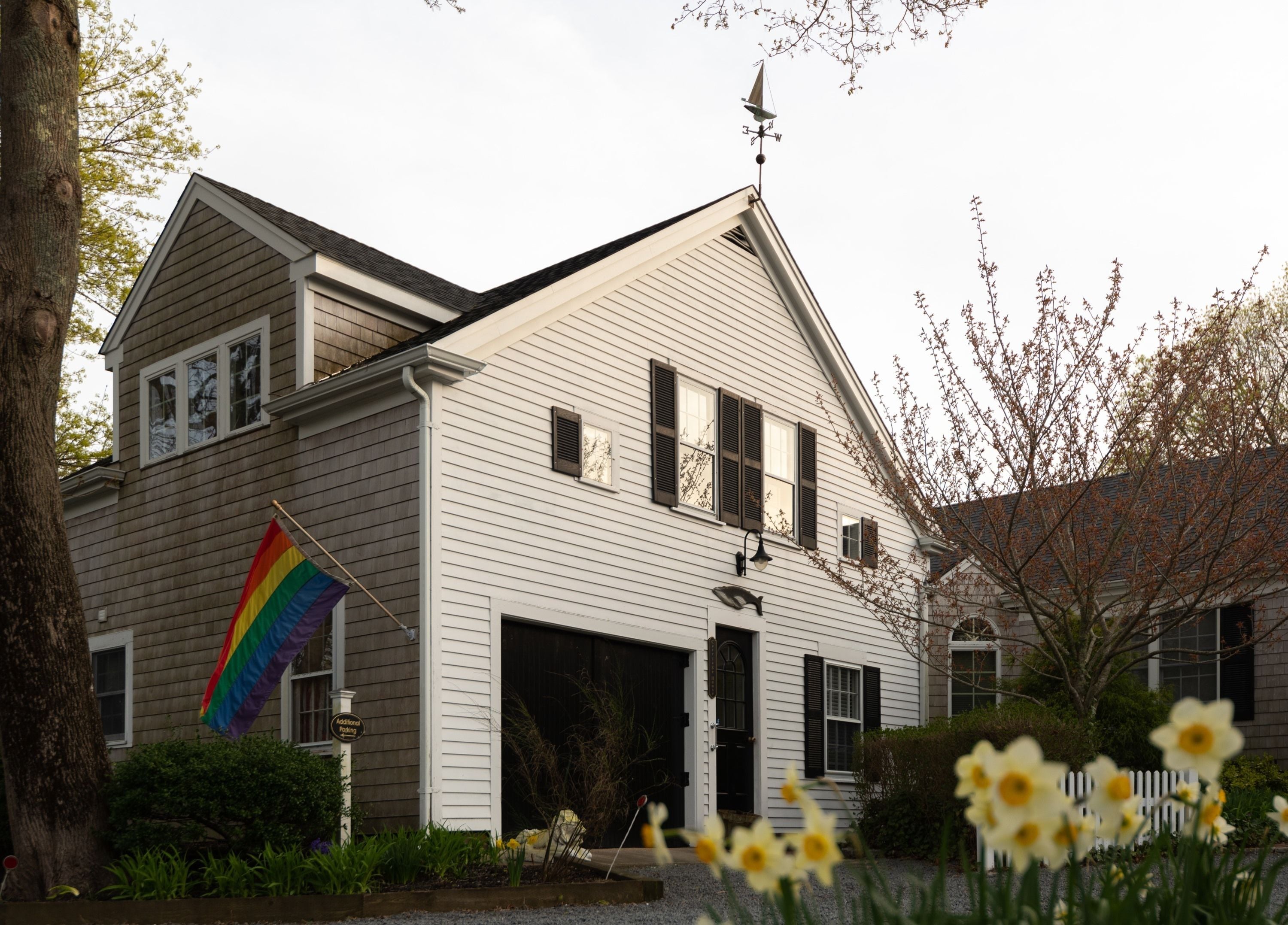 A two-story house with a garage, a flagpole with a rainbow flag, and daffodils in the foreground.